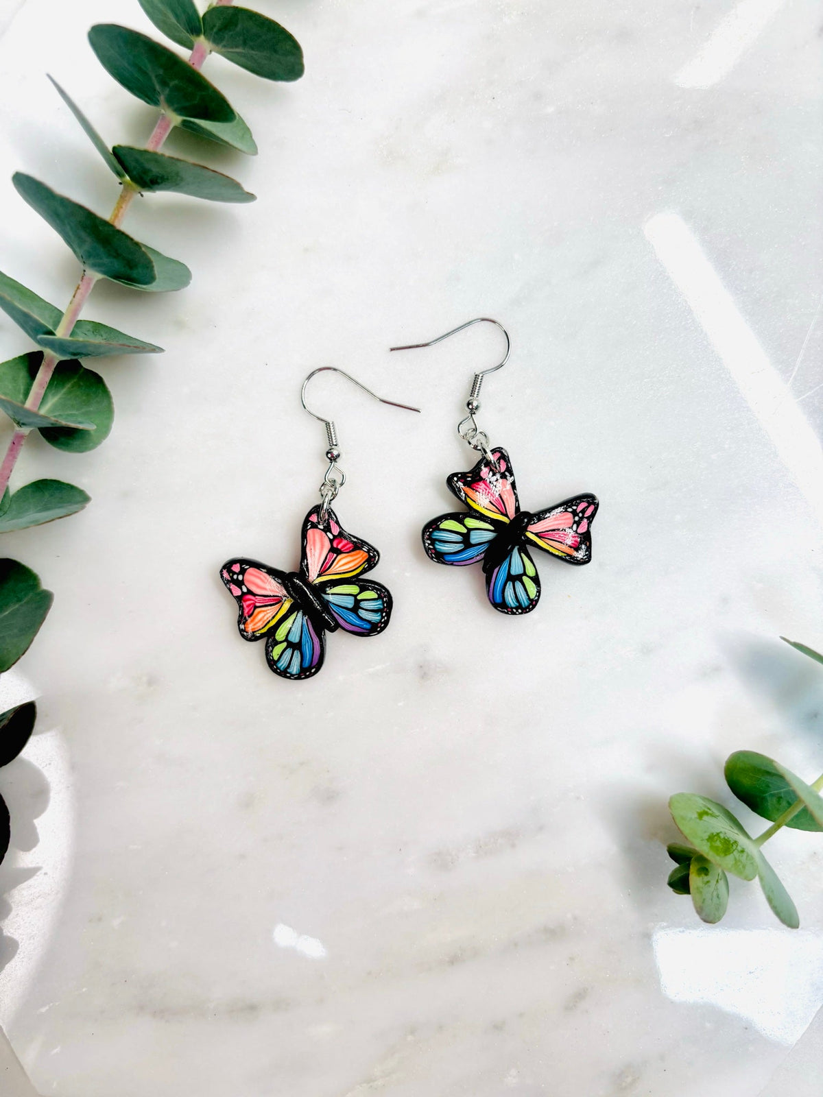 Rainbow butterfly earrings on marble with eucalyptus